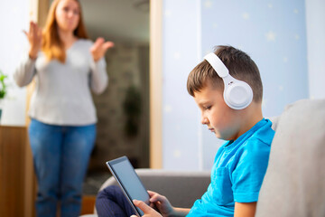 Mother scolds her son. Boy uses tablet with headphones and ignores his mom