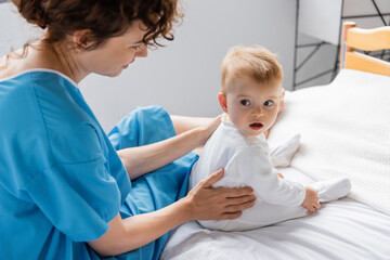 baby girl with open mouth looking away while sitting near mom on bed in clinic.