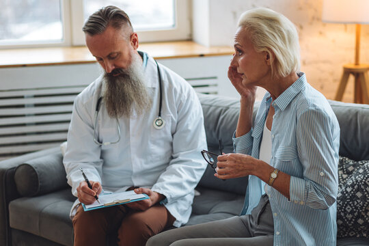 Male Doctor Visiting Attractive Senior Woman At Home For Consultation, Medical Service And Support, Geriatric Help, Prescribing Illness Treatment. Clinic Or Hospital Appointment For Eye Pain