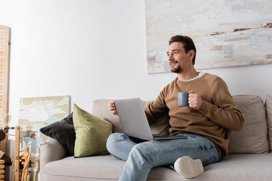 smiling man using laptop while holding cup and sitting on sofa in living room.