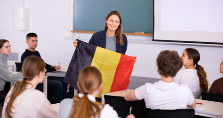Adult female teacher in the classroom showing the flag of Belgium to the students at the geography lesson