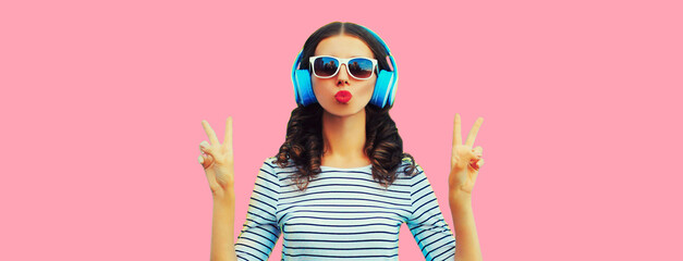 Portrait of stylish young woman in headphones listening to music blowing her lips sends sweet air kiss on pink background
