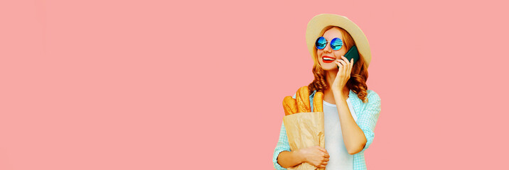 Portrait of smiling young woman calling on phone holding grocery shopping paper bag with long white bread baguette on pink background, blank copy space for advertising text