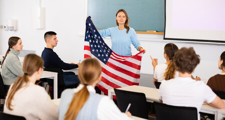 Geography lesson in school class - teacher talks about United States of America, holding a flag in...