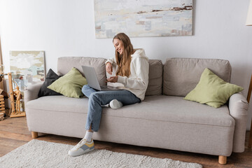 full length of positive woman in casual clothes having video call on laptop in living room.