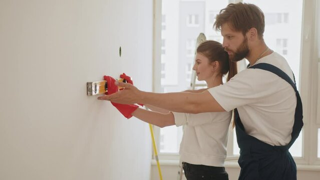 A happy family is renovating a purchased apartment. Smiling couple doing repair at home using spirit level to measure. Repair, building and home concept. Young woman using leveling tool at home