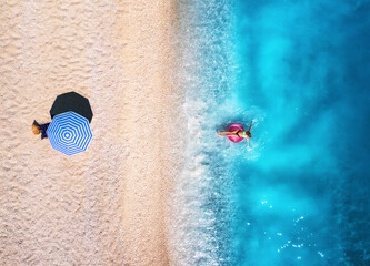 Aerial view of a woman swimming with pink swim ring in the sea, sandy beach and blue umbrella at...