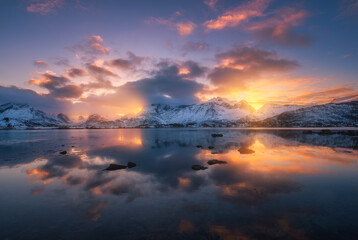 Sea coast, beautiful snowy mountains and colorful sky with clouds and golden sunlight at sunset in winter. Lofoten islands, Norway. Landscape, rocks in snow, reflection in water at dusk. Scenery - obrazy, fototapety, plakaty