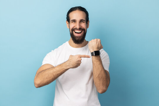 Look At Clock, No Rush. Portrait Of Man With Beard Wearing White T-shirt Pointing Wrist Watch And Expressing Optimism About Time, Not Busy. Indoor Studio Shot Isolated On Blue Background.