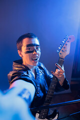 Obraz premium Punk rocker showing rock sign on camera and playing guitar, creating musical performance and acting crazy. Artist producing heavy metal alternative music with instrument, live performance.