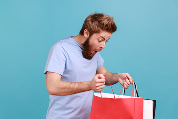 Portrait of attractive bearded man standing, holding red and white shopping bags and looking inside with shocked facial expression. Indoor studio shot isolated on blue background. - obrazy, fototapety, plakaty