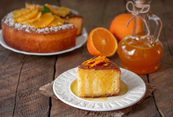 Slice of fluffy chiffon cake topped with orange zest strips on a plate. Bottle of orange syrup, fresh fruits and cake on background. Wooden table, selective focus, horizontal.