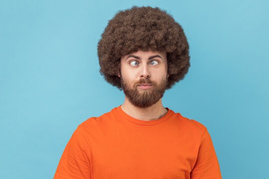 Portrait of crazy funny man with Afro hairstyle wearing orange T-shirt standing with crossed eyes and looking with silly comedian face. Indoor studio shot isolated on blue background.