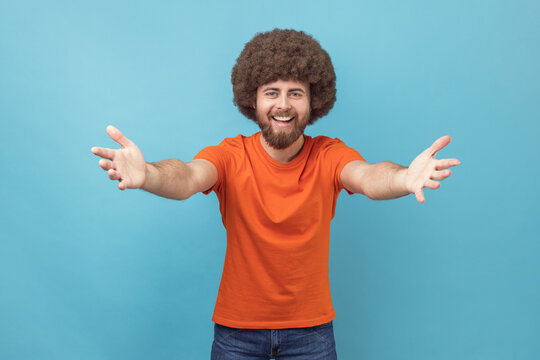 Come into my arms. Portrait of satisfied man with Afro hairstyle wearing orange T-shirt reaching out to camera, stretching arms to hug you. Indoor studio shot isolated on blue background.