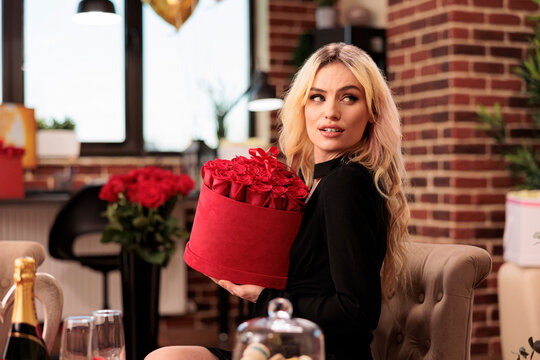 Lovely girlfriend holding valentines day red roses bouquet, receiving romantic gifts from boyfriend during love holdiday. Cute woman sitting in living room filled with macarons cookies and presents