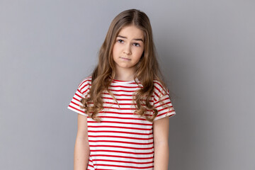 Obraz premium Portrait of unhappy little girl wearing striped T-shirt being upset of bad news, looking at camera with frowning face, expressing sadness. Indoor studio shot isolated on gray background.