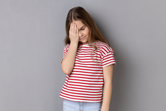 Oh no, I forgot. Portrait of little girl wearing striped T-shirt making facepalm gesture, feeling regret and shame about forgotten event, bad memory. Indoor studio shot isolated on gray background.