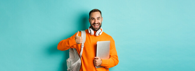 Happy man with backpack and headphones, holding laptop and smiling, showing thumb-up in approval,...
