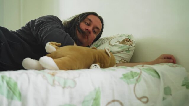 A guy with a plush toy is resting on a mattress lying on the floor. The guy has long hair