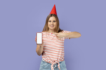 Portrait of smiling delighted blond woman in striped T-shirt and party cone holding and pointing at cell phone with empty screen for advertisement. Indoor studio shot isolated on purple background.