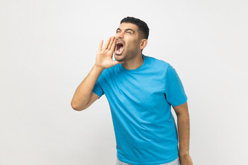 Portrait of excited handsome attractive unshaven man wearing blue T- shirt standing keeps hands near opened mouth, screaming news. Indoor studio shot isolated on gray background.