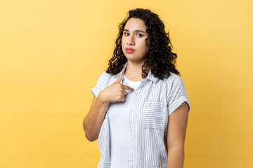 Portrait of serious self confident woman with dark wavy hair pointing at herself, feeling proud and self-important, having big ego. Indoor studio shot isolated on yellow background.