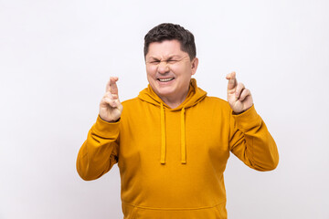 Fototapeta premium Portrait of middle aged man crossing fingers and closing eyes making wish, believing in miracle, hope in success, wearing urban style hoodie. Indoor studio shot isolated on white background.