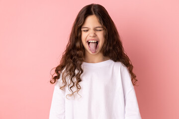 Portrait of childish carefree little girl wearing white T-shirt showing out tongue and closing eyes...