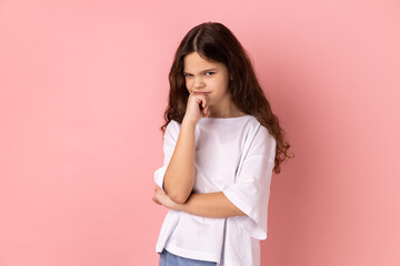 Need to think. Portrait of thoughtful little girl wearing white T-shirt holding her chin and pondering idea, confused not sure about solution. Indoor studio shot isolated on pink background.