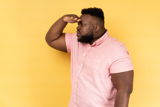 Side view of curious man with beard in pink shirt keeping palm over head and looking attentively far away, peering with expectation at long distance. Indoor studio shot isolated on yellow background.