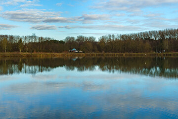Lake surface view with reflective trees and blue sky with white clouds. Autumn landscape.