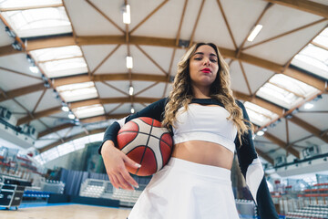 Low-angle shot of a blond cheerleader holding a basketball under her arm and squinting at the...
