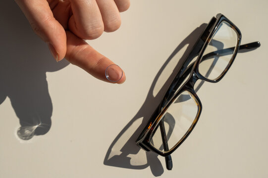 Close-up of a contact lens on a female index finger against the background of glasses on a white table. 