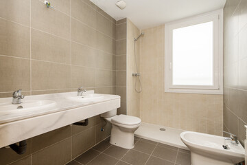 New mirrorless and partitionless bathroom fitted with white porcelain toilets and gray and cream tiles, window in shower and double sink over marble countertop