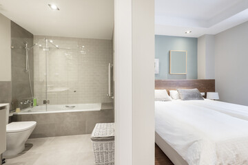 Bathroom with bathtub with glass partitions, gray tiles and bedroom with a double bed with a white duvet