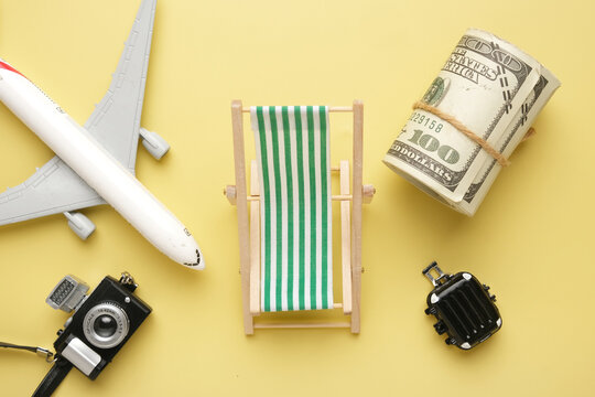 A flatlay picture of beach folding chair, roll of fake money, suitcase, toy camera and aeroplane on yellow background. Vacation plan concept.