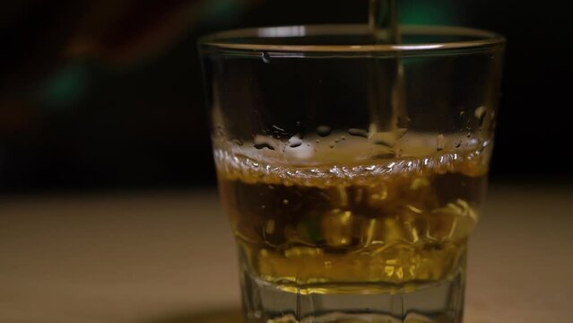Close up video of a person pouring whiskey into a shot glass. golden yellow drink is poured into a clear glass against a dimly lit background in the pub