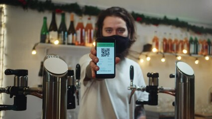 Barman in mask showing qr code. Bearded man behind bar counter holding covid vaccination certificate. Visiting pubs during coronavirus, green zone restrictions. Professional bartender in restaurant. - Powered by Adobe
