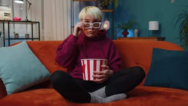 Excited woman with blonde hair sits on sofa eating popcorn snacks and watching interesting TV serial, sport game, film, online social media movie content at home. Girl enjoying evening entertainment