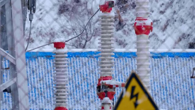 Transformer substation with wires, insulators, electric busbars, power transformer, switchgears, switching equipment in winter during snowfall. Lightning symbol in a yellow triangle - "high voltage" -