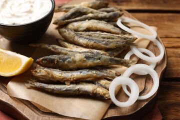 Delicious fried anchovies with onion rings, sauce and slice of lemon served on wooden table, closeup