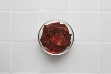 Bowl with pieces of delicious beef jerky on white tiled table, top view