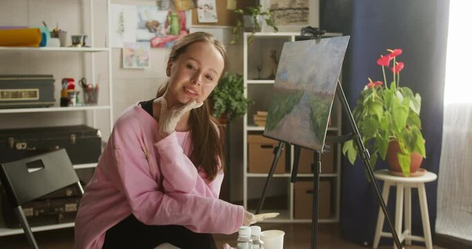 Young smiling woman artist portrait, painter wearing gloves sitting near beautiful picture landscape on easel in workshop. Female drawing teacher posing in art school, looking in camera.