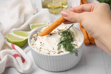 Woman dipping tasty grissini stick into cream cheese at white tiled table, closeup