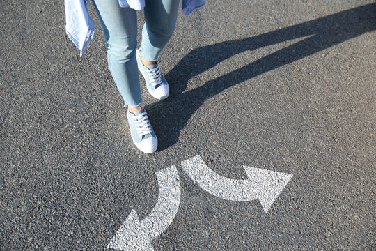 Choice Of Way. Woman Walking Towards Drawn Marks On Road, Closeup. White Arrows Pointing In Opposite Directions