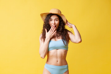 young shocked brunette girl in a blue swimsuit and with curly hair holds sunglasses on a yellow background
