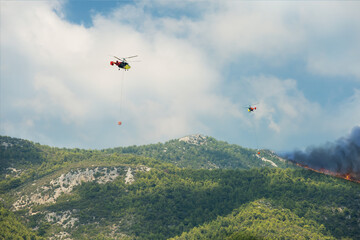 Two helicopters dumping water on forest fire in Mediterranean region.