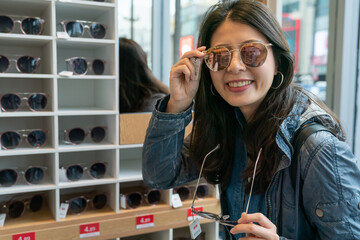 Smiling young asian girl traveler trying on sunglasses on mirror in optician face looking camera.