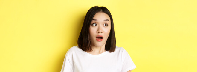 Close up of surprised asian girl drop jaw, gasping and looking left at logo, standing over yellow...