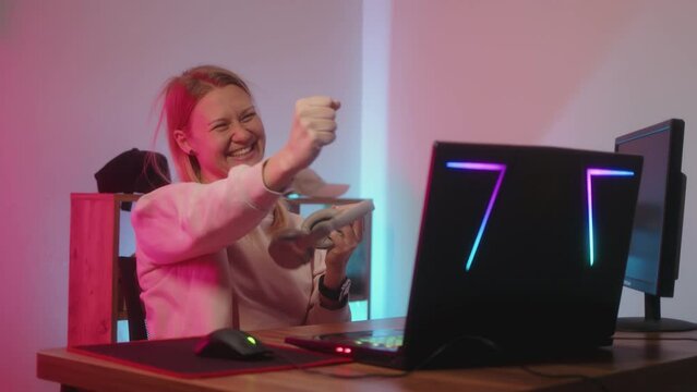 girl gamer plays on computer in purple-blue backlight of room and wins online match in game. young woman rejoices and raises her hands up in happiness. Winning difficult deathmatch.